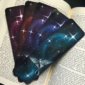 May include: A collection of five celestial-themed bookmarks. Each bookmark features a vibrant galaxy design with stars and a gradient of colors, with one showcasing a white moth illustration. The bookmarks are resting on an open book.