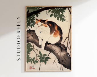 Op de afbeelding: Een ingelijste kunstdruk met een lapjeskat op een tak. De kat heeft witte, bruine en oranje vacht en draagt een blauwe halsband. Het kunstwerk is in traditionele Japanse stijl, met groene bladeren en een lichte achtergrond. De tekst "STUDIOFREELY" staat aan de linkerkant.