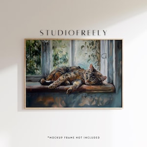 Op de afbeelding: Een ingelijst schilderij van een lapjeskat die op een vensterbank slaapt. De kat is bruin en oranje, en het raam biedt uitzicht op groen. Het schilderij zit in een houten lijst. De tekst "STUDIOFREELY" staat bovenaan.