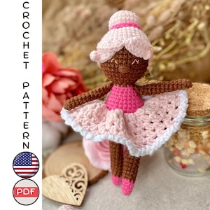 Puede incluir: Patrón de crochet para una muñeca bailarina con piel morena, vestido rosa y blanco y un moño rosa. La muñeca está sentada sobre una superficie de madera con un objeto en forma de corazón a su lado.