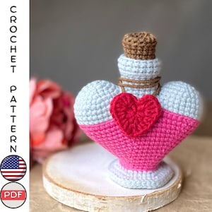 May include: Crochet pattern for a heart-shaped bottle with a cork stopper and a small red heart charm. The bottle is crocheted in light gray and pink yarn.
