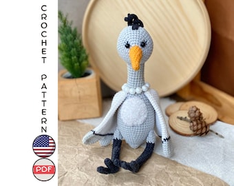 May include: A crocheted grey bird with black legs and a yellow beak. The bird is wearing a white pearl necklace and has black eyes. The bird is sitting on a brown surface.