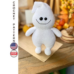 Könnte beinhalten: Ein weißes gehäkeltes Geister-Amigurumi-Spielzeug mit schwarzen Augen und einem lächelnden Mund. Der Geist steht auf einem braunen Papierbeutel auf einem Holztisch.