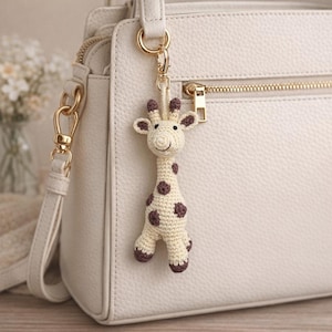 May include: A crocheted giraffe keychain with a cream-colored body and brown spots and accents. The giraffe is attached to a gold-colored metal clasp, hanging from a white handbag. The giraffe has a smiling face.