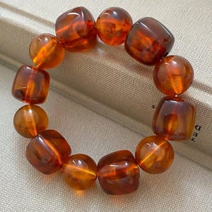 May include: A bracelet featuring alternating rounded and square amber-colored beads. The beads are strung together, creating a warm, earthy tone. The bracelet is displayed on a neutral background, highlighting the natural beauty of the amber.