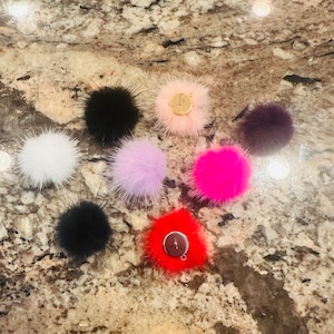 Puede incluir: Surtido de ocho pompones de piel sintética en varios colores, incluyendo negro, blanco, rosa, rojo, morado y gris. Cada pompon tiene un lazo de metal.