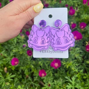 Puede incluir: Pendientes de acrílico iridiscente morado con forma de cabeza de dragón de dibujos animados. Los pendientes están en una tarjeta blanca con el texto "Don @KBRbyDonna".