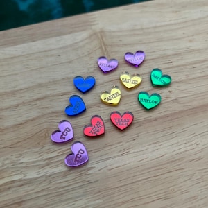 Könnte beinhalten: Eine Sammlung von 10 herzförmigen Acryl-Anhängern mit verschiedenen College-Namen und Farben. Die Anhänger sind auf einer Holzoberfläche angeordnet. Die Namen sind: Rhysand, Casteel, Baylor, April, TCU, Texas Tech und Day of.