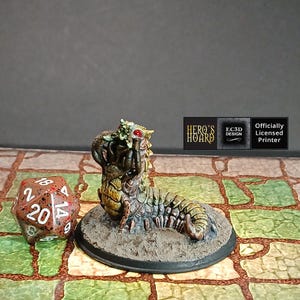 May include: A detailed miniature of a fantasy creature with a segmented body, textured carapace, and a menacing head with red eyes. The figure is mounted on a circular base. A twenty-sided die is next to the figure. The image includes the text "HERO'S HOARD".