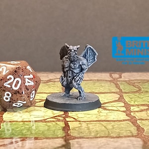 May include: A miniature gray gargoyle figurine with detailed wings and a menacing expression stands on a round base. A speckled brown twenty-sided die is next to the figurine. The background features a logo for Brite Minis.