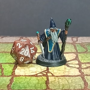 May include: A miniature wizard figurine with a long white beard, blue robes, and a pointed hat, holding a staff and a sword. Beside it is a large, speckled, twenty-sided die. The scene is set on a tiled surface.
