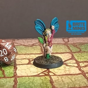 May include: A miniature fairy figurine with blue wings, a red top, and green boots, holding a staff with a green leaf. The figurine is on a circular base with a grassy texture. A twenty-sided die is in the background. The text "BRITE MINIS" is visible.