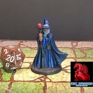 May include: A miniature wizard figurine in a blue robe and hat, holding a staff with a red orb. A twenty-sided die is next to the figurine. The Fat Dragon Games logo is in the lower right corner.