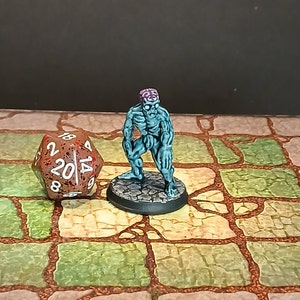 May include: A miniature zombie figurine with a teal-colored body and exposed brain. The figure stands on a circular base. A twenty-sided die with red and white markings is next to the figurine. The background is a stone-like surface.