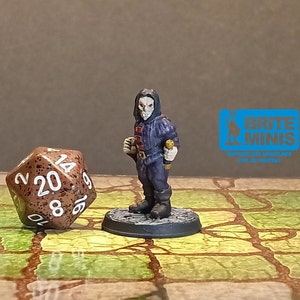 May include: A miniature figure of a robed character with a skull-like face, holding a small weapon. The figure is painted in shades of blue, red, and gray, standing on a circular base. A twenty-sided die is next to the figure.