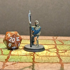 May include: A miniature skeleton warrior figurine with a spear, painted in blue and bone colors, stands on a circular base. Beside it is a twenty-sided die with brown and white markings. The scene is set on a stone-like surface.