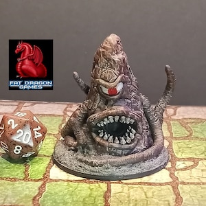 May include: A detailed, hand-painted miniature monster with a large open mouth, sharp teeth, and a single red eye. The creature is brown and gray, with textured skin. A twenty-sided die and the "Fat Dragon Games" logo are also visible.