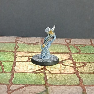May include: Miniature fantasy figure of a hammer-wielding character. The figure is primarily silver and gold, with a blue, smoky base. It stands on a black, circular base, and is set against a backdrop of a stone floor.