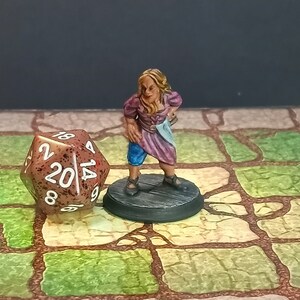 May include: A miniature figurine of a female character in a purple and blue outfit, standing on a wooden base. A large, brown twenty-sided die is next to the figure. The scene is set on a stone-patterned surface, suggesting a tabletop game.