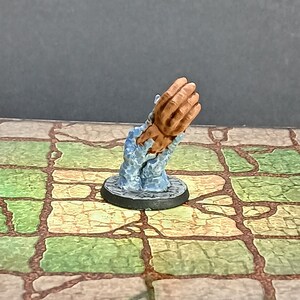 May include: A miniature figurine of a pair of hands emerging from a blue, textured base. The hands are painted a flesh tone, with detailed fingers. The base is round and black, and the figurine is likely for tabletop gaming or display.