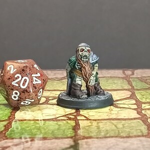 May include: A miniature fantasy figure of a skeletal dwarf with a long brown beard, green tunic, and metal shoulder armor. A twenty-sided die with brown and red speckles is next to the figure. The miniature is on a round base.