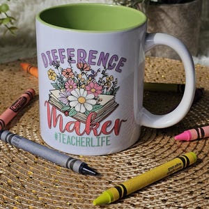 Peut inclure: Mug en céramique blanche avec intérieur vert, avec les mots "DIFFERENCE Maker" et "#TEACHERLIFE" avec un motif floral. Des crayons Crayola de différentes couleurs entourent la tasse sur une surface tissée.