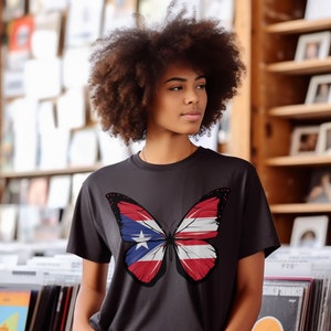 Puede incluir: Una persona lleva una camiseta gris oscuro con un diseño de mariposa. Las alas de la mariposa están decoradas con los colores de la bandera puertorriqueña.
