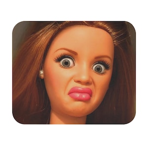 May include: A doll head with long brown hair, wide grey eyes, long black eyelashes, and bright pink lips. The doll has a facial expression with a furrowed brow and pursed lips, and a small silver-colored stud earring is visible on the left earlobe.