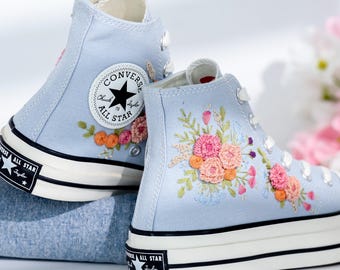 May include: A pair of light blue high-top Converse sneakers with embroidered floral designs. The sneakers have black laces and a white rubber sole. The Converse logo is visible on the side of the sneakers.