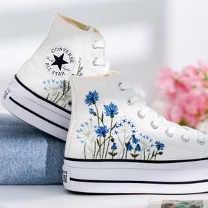 May include: White high-top Converse sneakers with a platform sole and blue floral embroidery. The Converse All Star logo is visible on the side of the shoe.