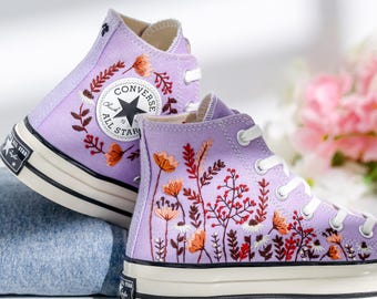 May include: Purple high-top Converse sneakers with embroidered floral designs. The sneakers have a white rubber sole and a black star logo.