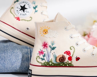 May include: White high-top Converse sneakers with embroidered flowers, a snail, a mushroom, and a ladybug. The sneakers also feature a yellow sun with a smiling face and a white cloud.