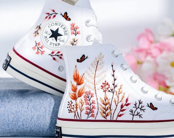 May include: White high-top Converse sneakers with embroidered floral designs and butterflies. The sneakers have a red stripe around the sole and the Converse All Star logo on the side.