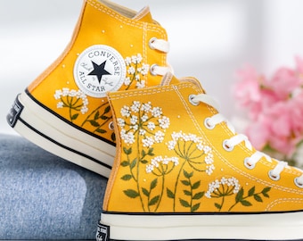 May include: Yellow high-top Converse sneakers with white embroidered flowers and green leaves. The Converse logo is visible on the side of the shoe.