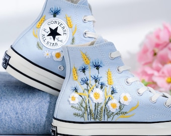 May include: Light blue high-top Converse sneakers with embroidered flowers and wheat stalks. The Converse All Star logo is visible on the side of the shoe.