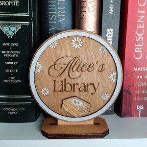 May include: A wooden sign with a white border and a white background. The sign has a brown wooden base and says "Alice's Library" in brown lettering. The sign is decorated with white daisies and a stack of books.