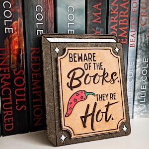 Peut inclure: Une enseigne en bois rectangulaire en forme de livre avec une bordure marron foncé et un centre beige clair. Le texte indique : Beware of the Books. They're Hot. Un petit piment rouge à motifs d'étoiles est imprimé sous le mot Books. Quatre étoiles blanches décorent les coins.