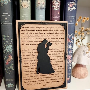 Peut inclure: Une œuvre d'art encadrée avec une silhouette noire d'un couple enlacé, sur fond de texte manuscrit. L'œuvre est exposée sur une étagère avec des livres anciens, dont des titres comme "Jane Austen" et "Charlotte Brontë."