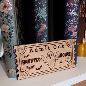 Könnte beinhalten: Holzschild "Admit One Haunted House" mit Geister-, Fledermaus- und Spinnendesign. Das rechteckige Schild steht vor alten Büchern.