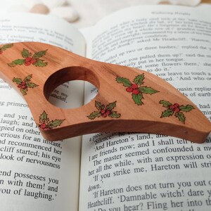 May include: A wooden bottle stopper with a holly design. The stopper is brown with green holly leaves and red berries. The stopper is sitting on top of an open book.