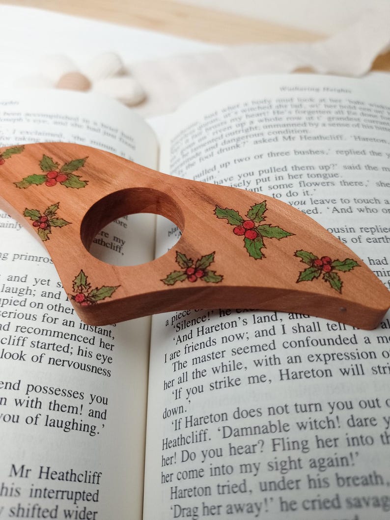 May include: A wooden bottle stopper with a holly design. The stopper is brown with green holly leaves and red berries. The stopper is sitting on top of an open book.