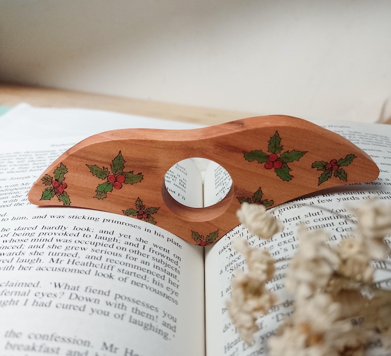 May include: A wooden bookmark with a holly design. The bookmark is shaped like a wave and has a hole in the center. The holly leaves are green and the berries are red.