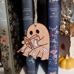 May include: A wooden ghost-shaped bookmark reading a book with "Autumn" on the cover, with a pumpkin and leaves design. The bookmark hangs in front of a row of vintage books with floral designs and the titles "Edith Wharton" and "Jane Eyre".