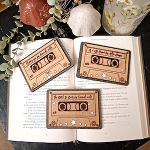 Peut inclure: Trois ornements rectangulaires en bois en forme de cassettes audio, chacun présentant des textes gravés et des icônes décoratives. En haut à gauche, Cherry pie by hannah wells, en haut à droite, U + Ur Hand by Allie Hayes, et en bas, The Bitch Is Back by hannah wells.