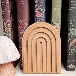 Puede incluir: Un sujetalibros de madera en forma de arcoíris con acabado natural se encuentra frente a una fila de libros antiguos. El sujetalibros tiene un diseño en capas con múltiples arcos. Los libros tienen cubiertas florales y decorativas.