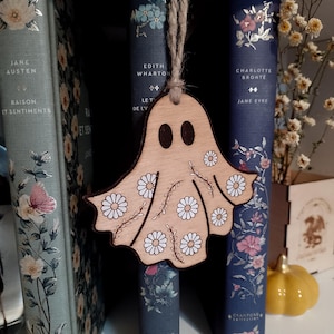 Può includere: Un ornamento in legno a forma di fantasma con dettagli a margherita, appeso davanti a una libreria. Il fantasma ha due fori per gli occhi e motivi a margherita sul corpo. Sono visibili libri con disegni floreali e titoli come "Jane Austen" e "Charlotte Brontë".