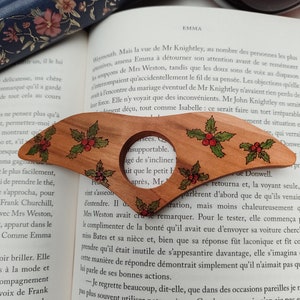 May include: A wooden bookmark with a holly design. The bookmark is shaped like a Christmas tree and has a red and green holly design with a red berry. The bookmark is resting on a page of a book with text about the novel Emma by Jane Austen.