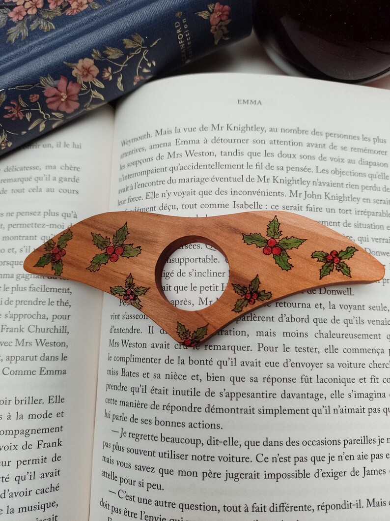 May include: A wooden bookmark with a holly design. The bookmark is shaped like a Christmas tree and has a red and green holly design with a red berry. The bookmark is resting on a page of a book with text about the novel Emma by Jane Austen.