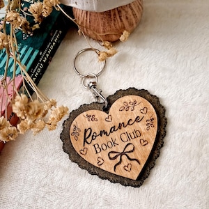Puede incluir: Un llavero de madera en forma de corazón con la inscripción "Romance Book Club". El llavero tiene un borde festoneado marrón oscuro y un llavero plateado. También incluye pequeños corazones y detalles florales.