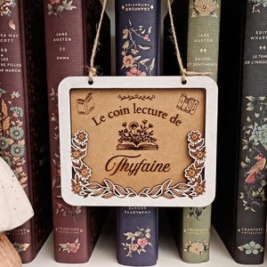 Puede incluir: Un letrero de madera con el texto "Le coin lecture de Thyfaine" cuelga frente a una fila de libros. El letrero tiene un marco blanco y un centro marrón con ilustraciones florales y de libros.